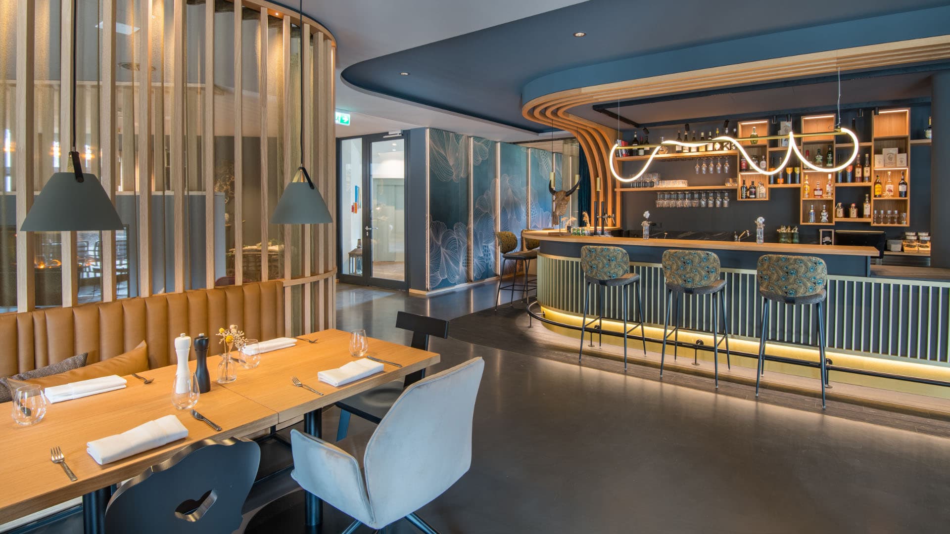 Innenarchitektur München / Innenarchitektur Federleicht - Offener Bar- und Speisebereich im La Soa Chalets mit geschwungenem Tresen, Holzlamellen und moderner Beleuchtung