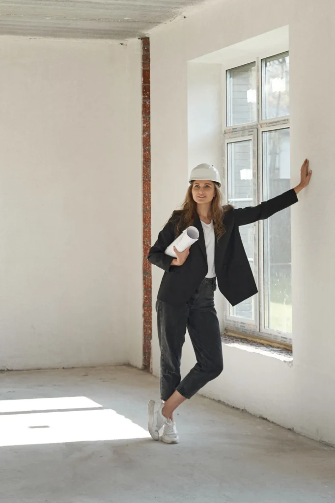 Architektin mit Bauhelm und Plänen steht lächelnd am Fenster eines Rohbaus